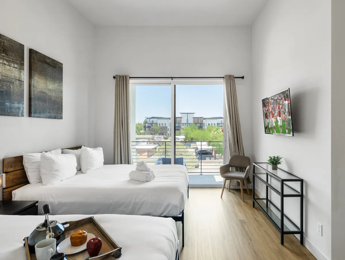 A modern bedroom with two Queen beds dressed in white linens, a large sliding glass door leading to a balcony, a wall-mounted TV for entertainment, and a small chair for relaxing or reading. Natural light fills the space, creating a welcoming, restful atmosphere.