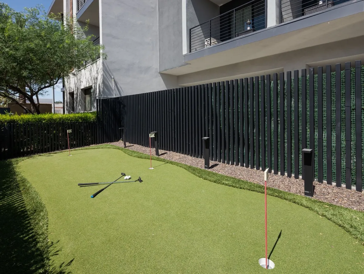 A well-manicured outdoor putting green bordered by modern architecture, a fence, and landscaped hedges. The space is bathed in natural light, evoking a sense of leisure and offering a dedicated spot for friendly golf practice or casual outdoor games.