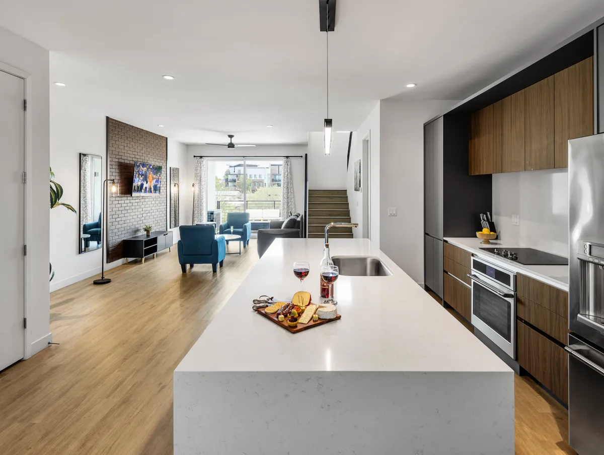 This image presents a welcoming, contemporary open-concept space, with a kitchen island adorned with a charcuterie board, blue chairs in the connected living room, and large windows that fill the area with natural light. The scene visually connects the kitchen and living area, highlighting spaces for cooking, dining, and socializing.