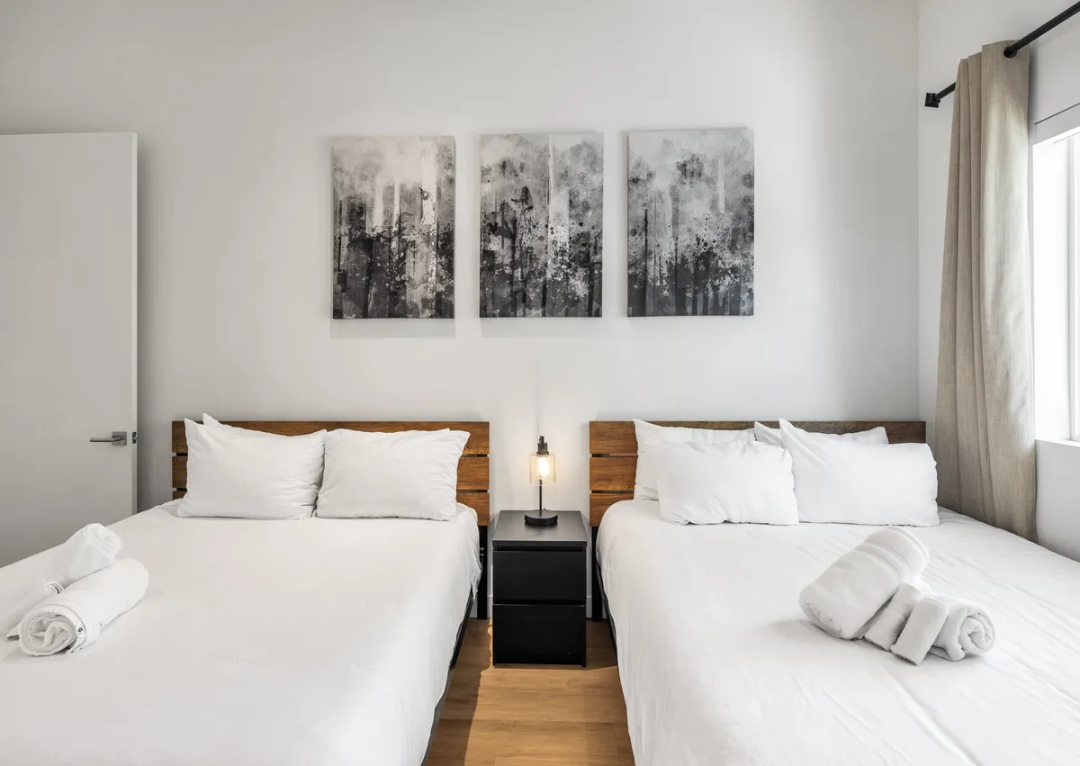 This image depicts a serene bedroom with two beds featuring crisp white linens, abstract artwork on the wall, and a nightstand with a softly glowing lamp. Natural morning light streams through the window, creating a relaxed and inviting atmosphere.