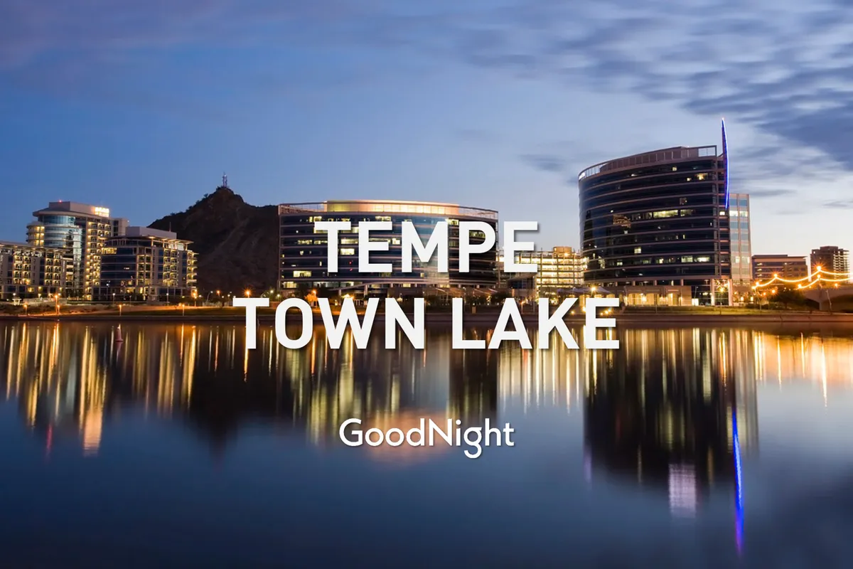 An exterior view of sleek, modern buildings reflected in a calm lake, with a dramatic mountain silhouette in the background. The setting sun casts warm hues over the scene, suggesting evening strolls, festivals, and tranquil moments by the water.