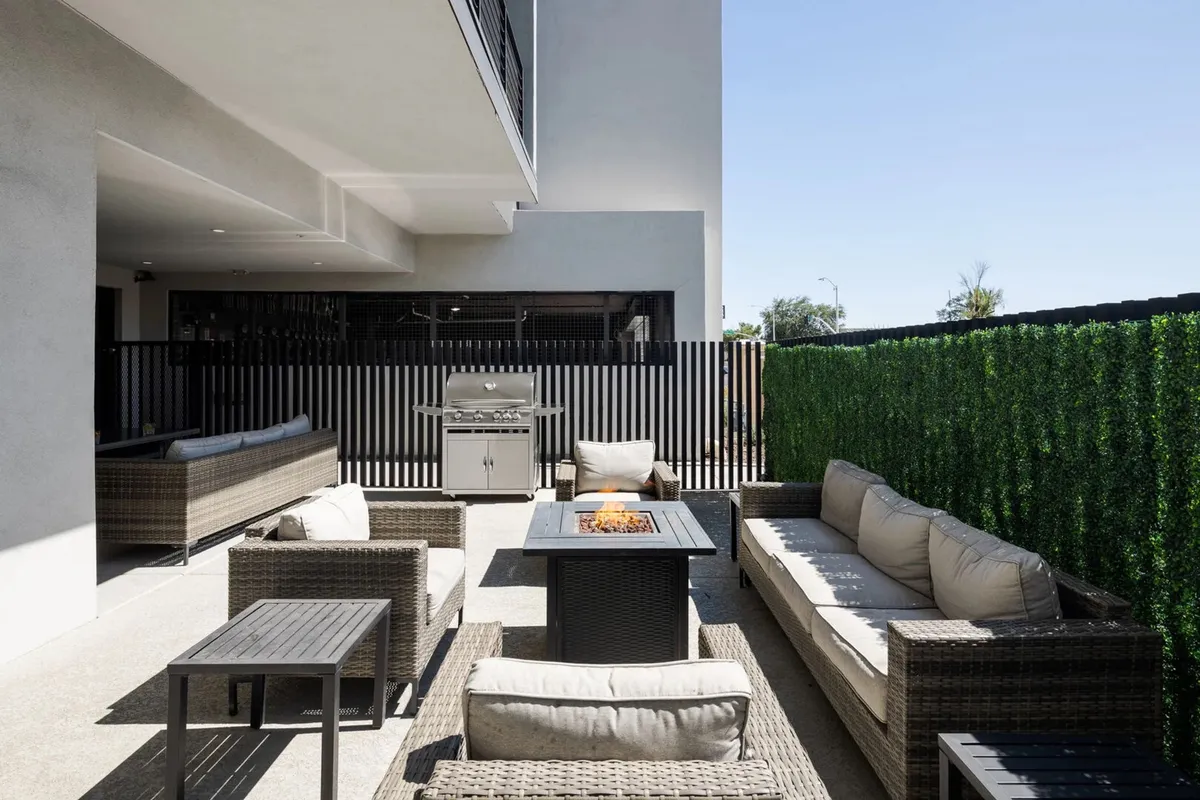 A modern outdoor setup featuring a fire pit at the center, surrounded by comfortable seating and a sleek barbecue grill. The area is enclosed with a greenery fence, creating a private yet inviting space perfect for evening gatherings, grilling, and sharing stories by the fire.