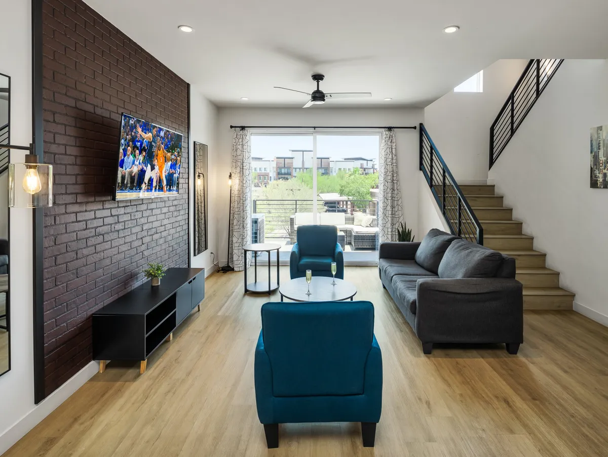 The image showcases a stylish living area with a striking brick accent wall, modern blue chairs, a large TV, and an open staircase, all arranged in a bright, inviting space ideal for relaxing or socializing.
