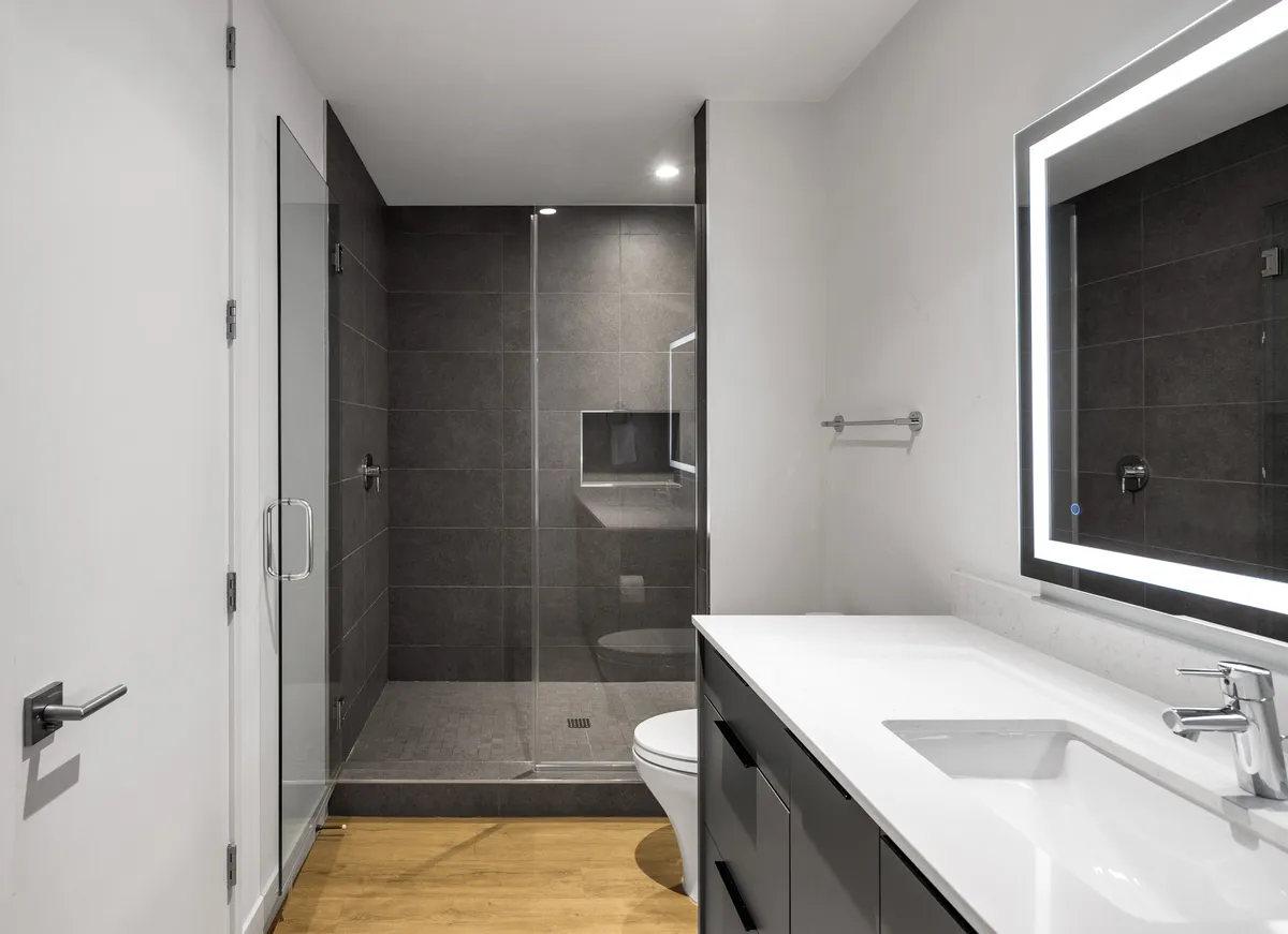 A serene, spa-inspired en-suite bathroom featuring a modern glass shower enclosure, a large backlit mirror, and a minimalist vanity. The space is bathed in soft morning light, evoking a sense of tranquility and rejuvenation.