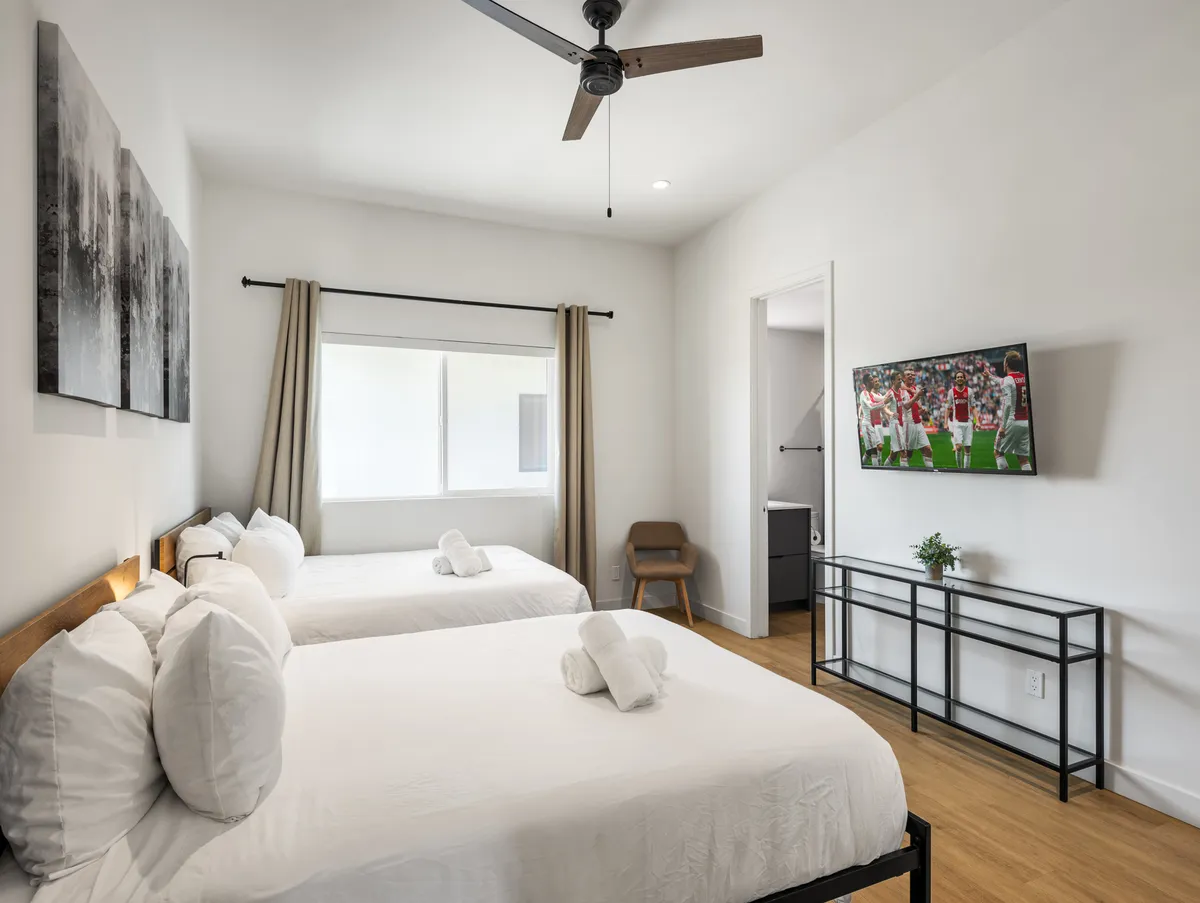 A stylish bedroom with two Queen beds, a wall-mounted smart TV, modern artwork, and a window with curtains for fresh air and privacy. The space feels bright and contemporary, perfect for recharging after a long day.
