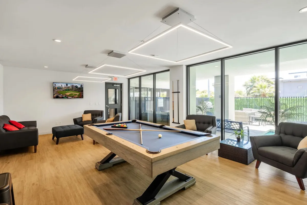 A vibrant clubhouse interior featuring a pool table as the centerpiece, surrounded by comfortable seating and large windows that let in plenty of natural light. A television is mounted on the wall, creating a lively, social atmosphere ideal for friendly games, watching sports, and relaxing with friends.