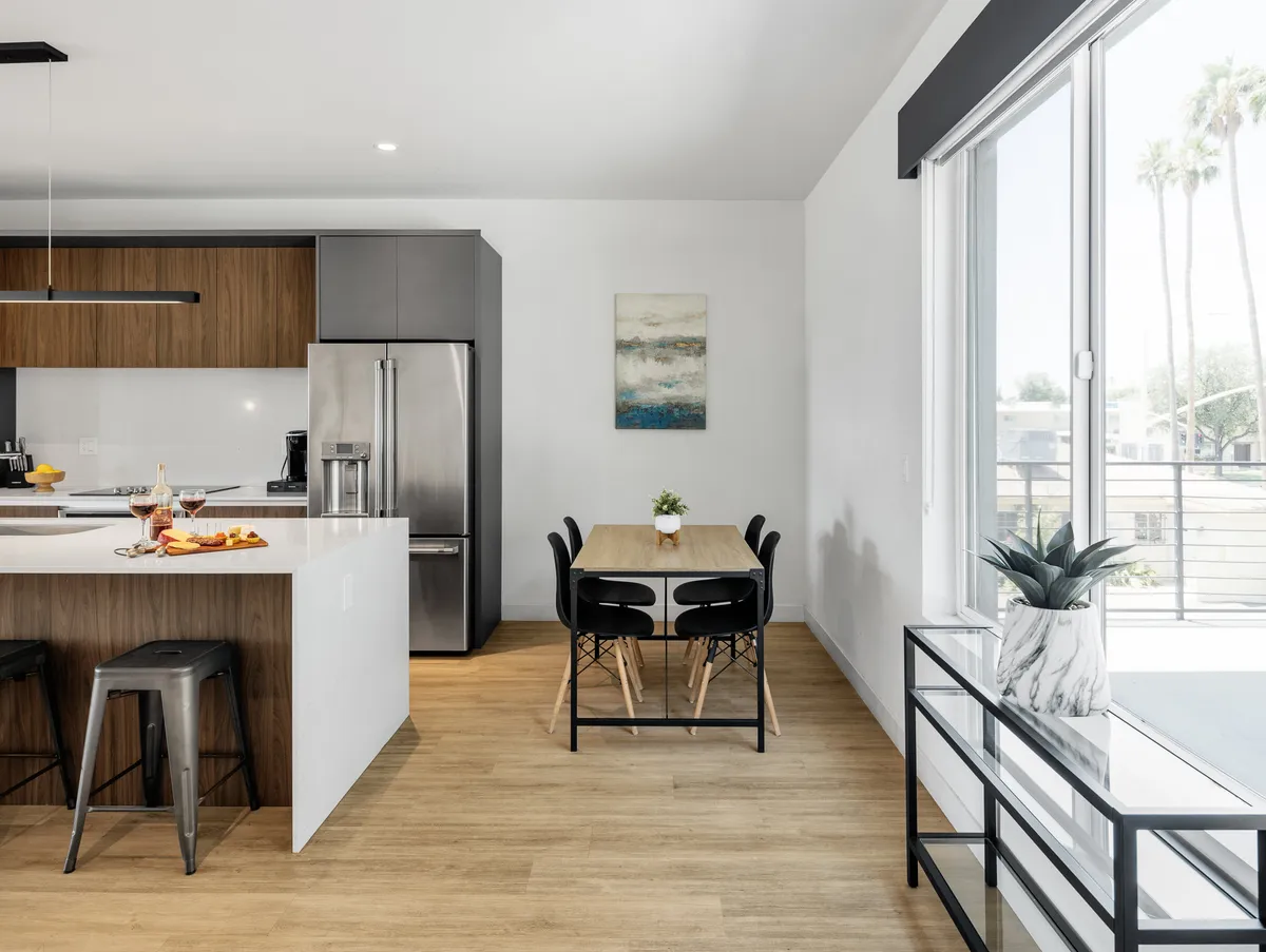 The image shows an open-concept kitchen and dining area with a modern kitchen island, sleek appliances, a dining table, and large windows that let in plenty of sunlight. The space feels both contemporary and welcoming, ideal for socializing and enjoying meals together.